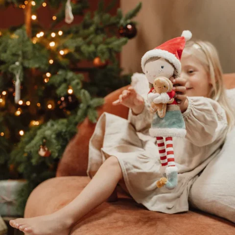 ενα κοριτσάκι κάθεται στο σαλόνι με το Χριστουγεννιάτικο δέντρο δίπλα και κρατάει την κούκλα με το κόκκινο σκουφάκι τις ριγέ κάλτσες και κρατάει έναν μπισκοτούλι